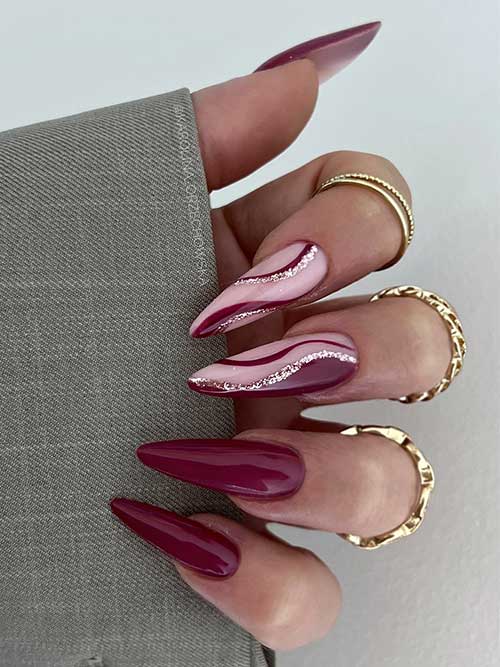 Long almond burgundy fall nails with two nude pink accent nails decorated with burgundy and rose gold swirls.