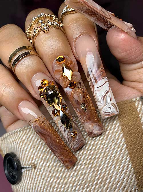 Long square-shaped brown and white marble nails embellished with glitter and rhinestones for a creative look.