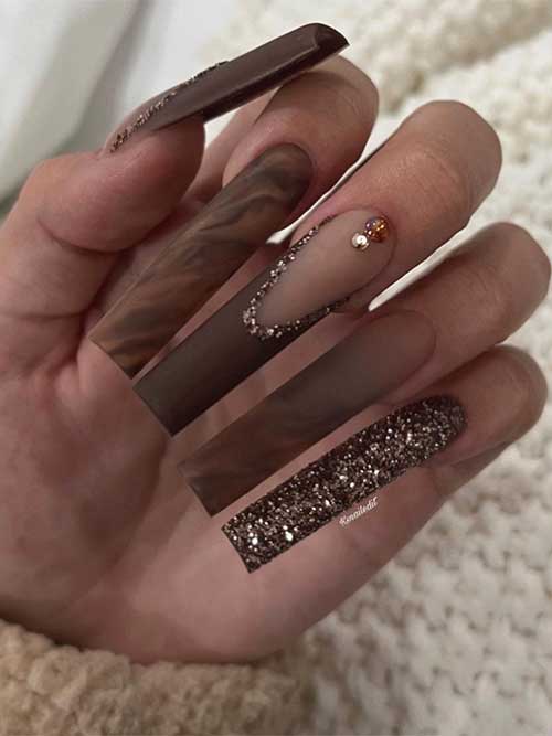 Long square-shaped matte dark brown nails with French tips adorned with gold glitter, rhinestones, and marble effect on two accent nails.