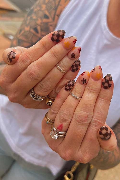 Short almond-shaped nude and brown checkered nails with red, brown, and burnt yellow flowers on two accent nude nails.