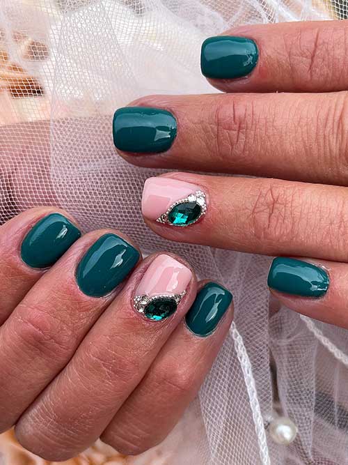 Short dark green nails with a nude accent featuring an emerald gem and silver rhinestones for a chic, elegant look.