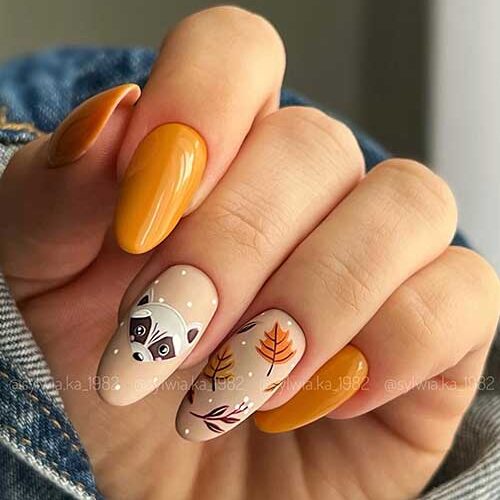 Burnt yellow Thanksgiving nails with two beige accent nails one of them adorned with fall leaves and the other fox nail art