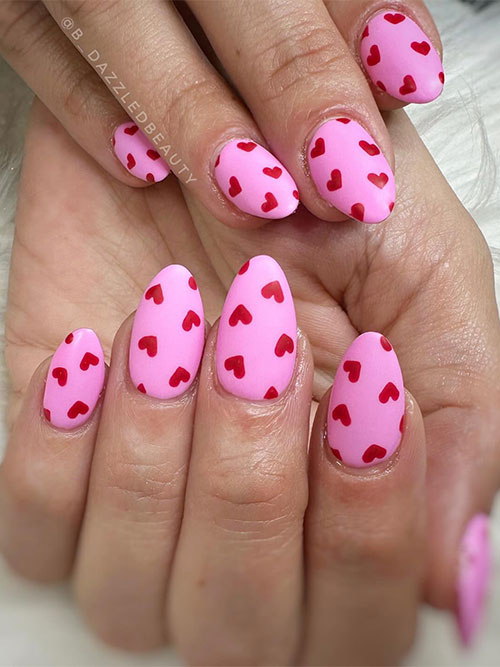 Short almond-shaped matte pink Valentine's Day nails adorned with tiny red heart shapes