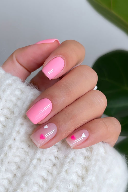 Short square-shaped pink Valentine's Day nails with two accent nude nails adorned with two white and hot pink heart shapes