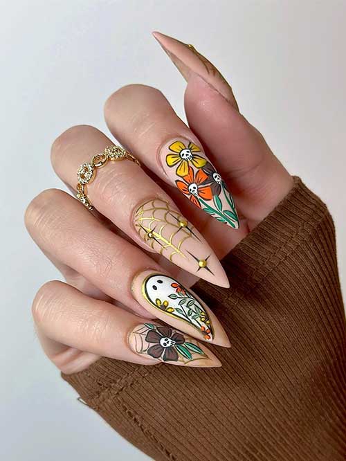 Matte stiletto nude Halloween nails decorated with autumnal colorful skull flowers, gold spider web, and a ghost with flowers