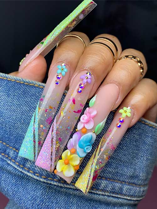 Long square nails in clear and pastel colors, adorned with vibrant 3D flowers and glittery accents for a springtime look.