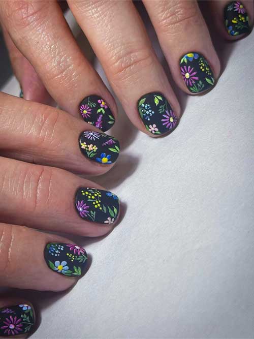 Matte black nails adorned with vibrant floral patterns in pink, yellow, blue, and white, accented by green leaves and stems.