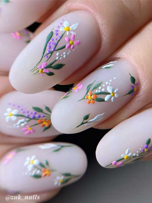 Matte light beige nails adorned with tiny white, pink, orange, and purple flowers, complemented by green leaves and white dots.