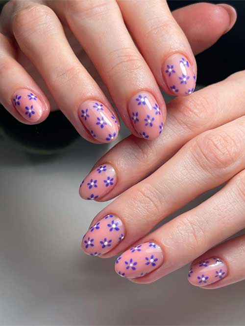 Soft nude pink nails adorned with delicate blue flowers, each featuring a darker blue center for a whimsical touch.